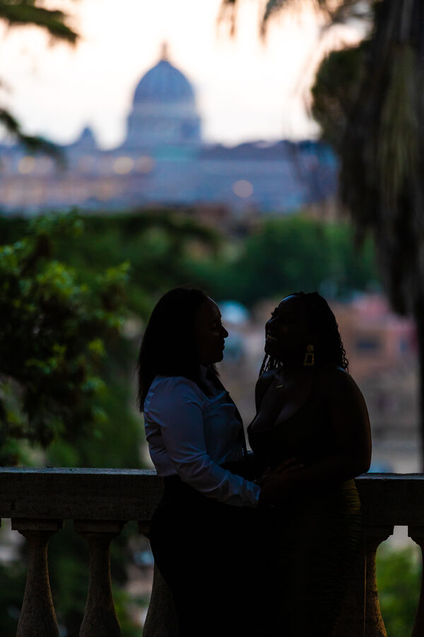 Low-key image of female gay couple holding each other at the Pincio Gardens with the Vatican in the background in the blue hour